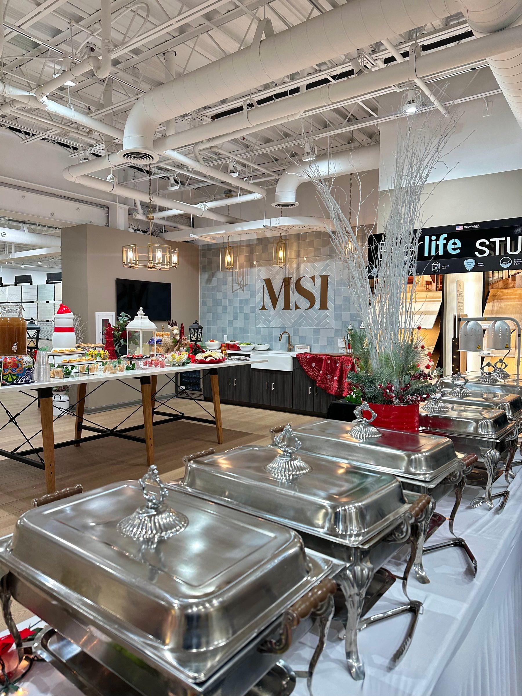 Silver chafing dishes lined up for a Gaelos corporate catering event in a showroom, with a holiday dessert table behind