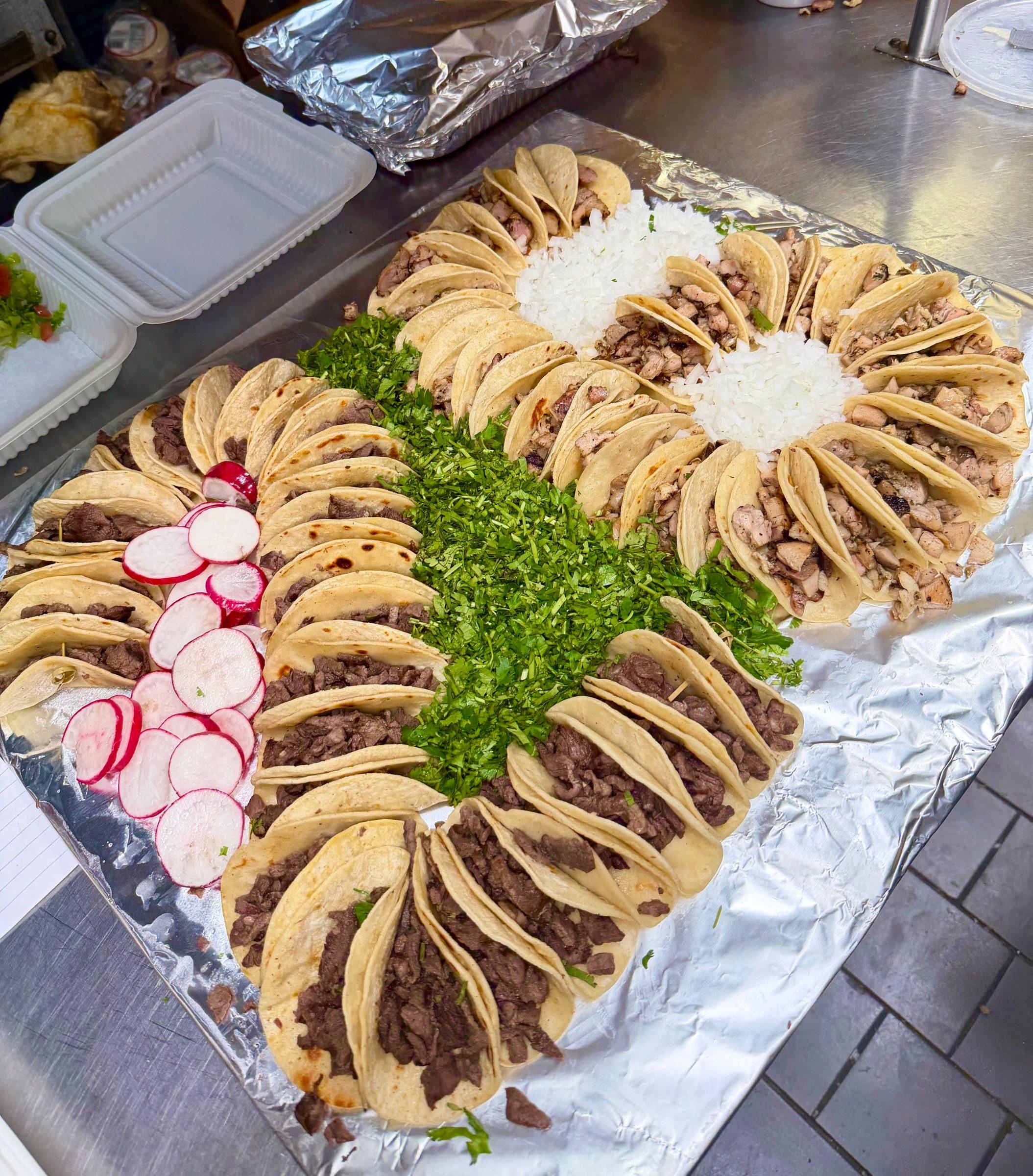 A large foil tray of street tacos laid out in rows around chopped cilantro, diced onion and sliced radishes
