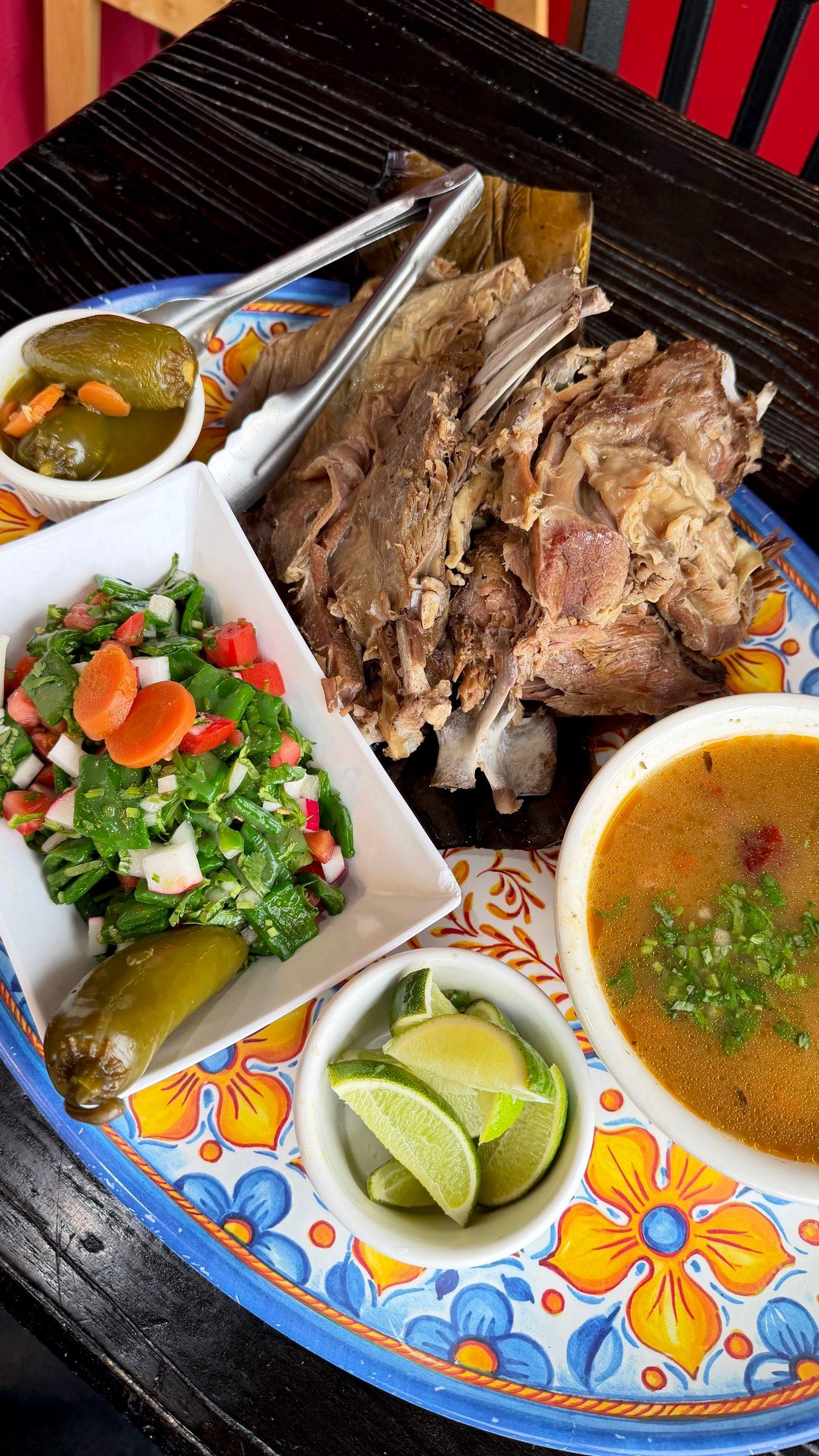 A Sunday barbacoa platter: slow-cooked barbacoa with a cup of consomé, nopal salad, pickled jalapeños and lime on a painted platter